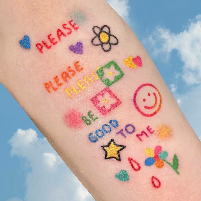 Colorful self-love tattoo with hearts, flowers, and the words "Please be good to me" on skin against a blue sky background.