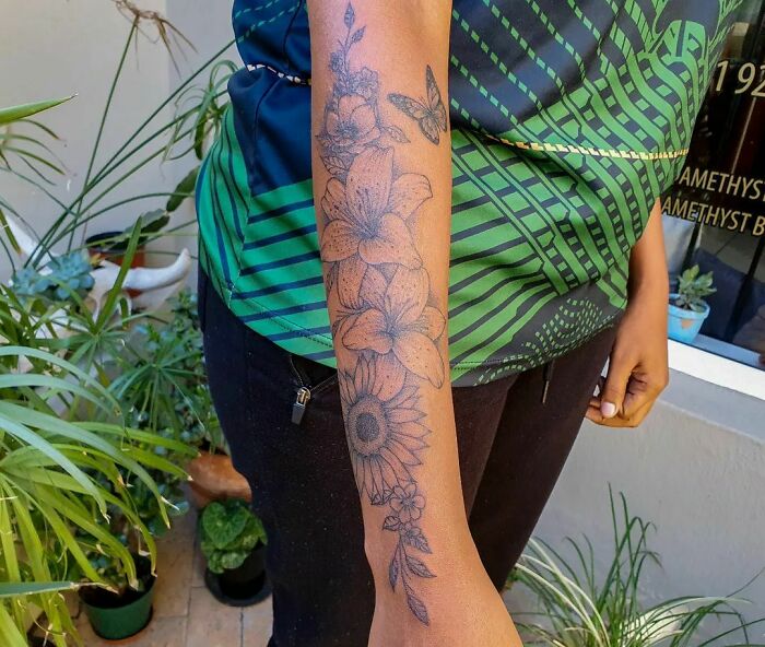 Arm adorned with detailed flower tattoo, showcasing elegance and beauty among lush green plants.