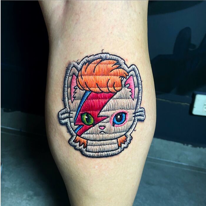 Tattoo by Eduardo Lozano, showcasing a colorful embroidered cat design on a person's leg.