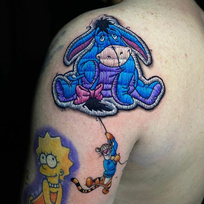 Tattoo art by Eduardo Lozano showing cartoon characters in a colorful, embroidery style on an arm.