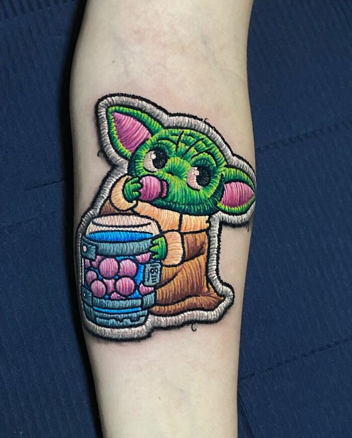 Tattoo of a cute green character sipping a drink, showcasing Eduardo Lozano's unique embroidery style.