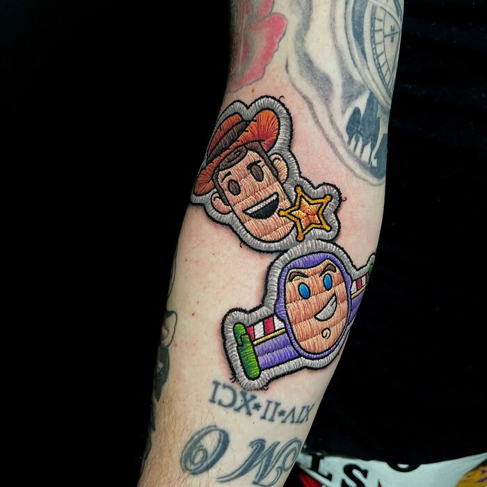 Embroidered-style tattoo by Eduardo Lozano featuring cartoon characters Woody and Buzz on a person's forearm.