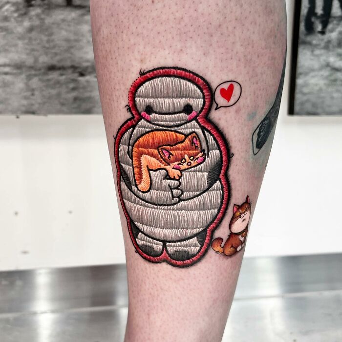 Tattoo by Eduardo Lozano featuring embroidered-style cat design with heart detail on leg.