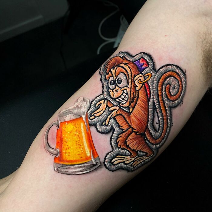 Tattoo of a cartoon monkey reaching for a frothy beer mug, showcasing a unique 3D effect by Eduardo Lozano.