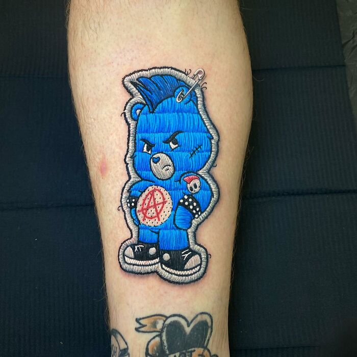 Embroidered-style tattoo of a blue bear with punk details on a person's leg, showcasing a new dimension in tattooing.