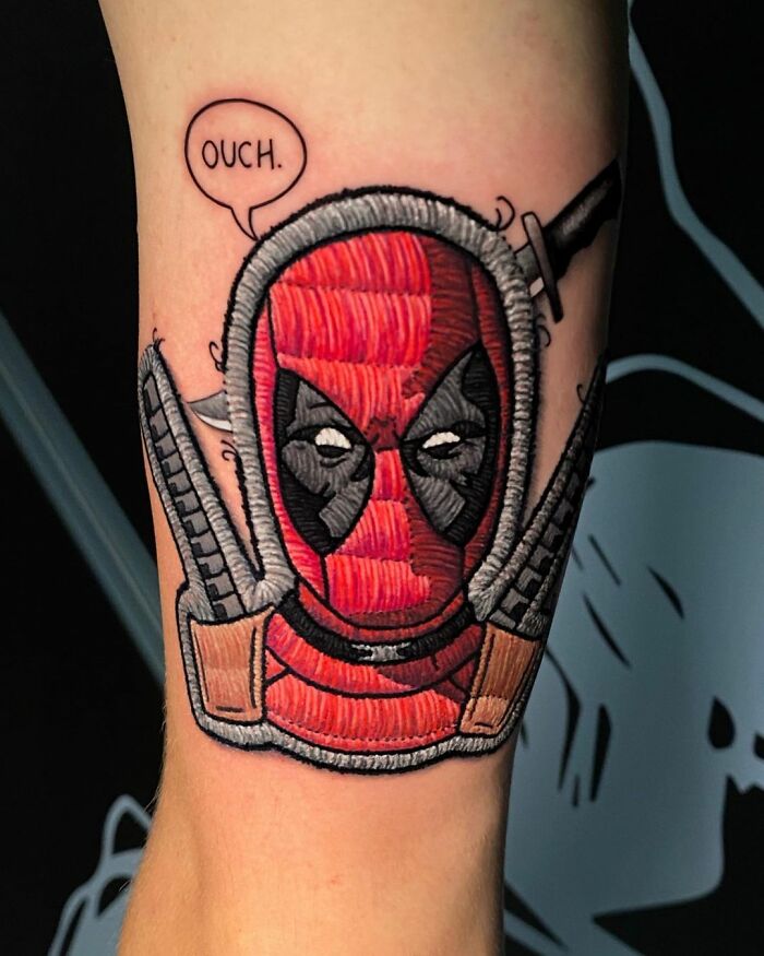 Tattoo of a 3D embroidered Deadpool character by Eduardo Lozano, featuring red and black design with swords and a speech bubble saying "Ouch."