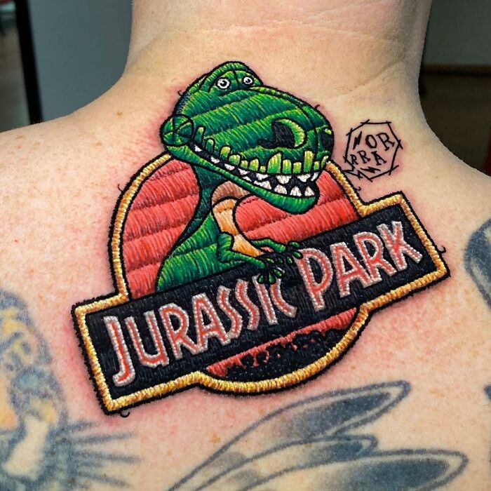 Tattoo of Jurassic Park logo with embroidered style by Eduardo Lozano on the back of a person's neck.
