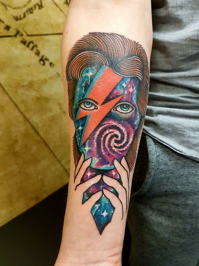Space tattoo featuring a colorful cosmic face with red lightning bolt and hands holding a galaxy pattern.