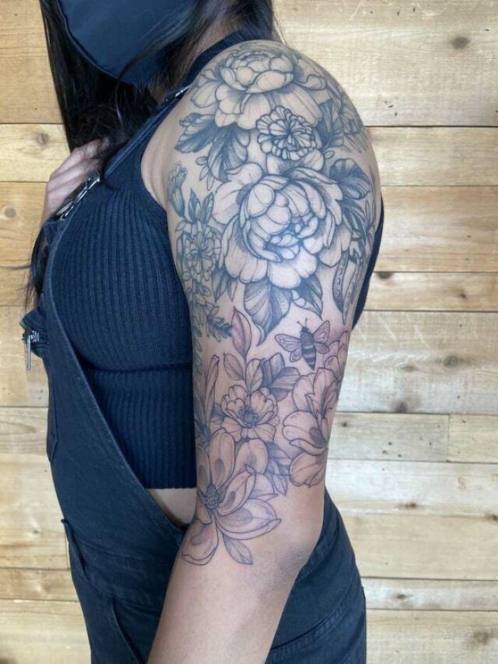 Floral tattoo on upper arm, featuring intricate blossoms and a bee, against a wooden background.