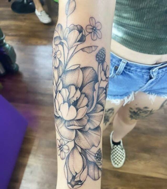 Flower tattoo design on forearm, featuring detailed blossoms and leaves, worn by someone in denim shorts and checkered shoes.