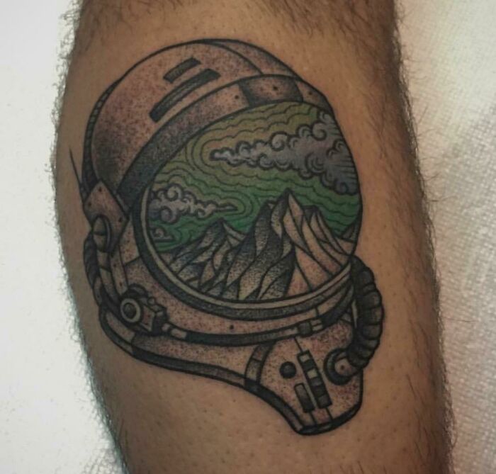 Space tattoo featuring a helmet with a scenic landscape inside, including mountains and clouds, on a person's arm.