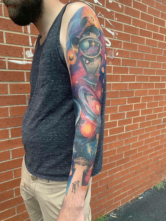 Man with colorful space tattoo sleeve, featuring planets and astronaut, standing against a brick wall.