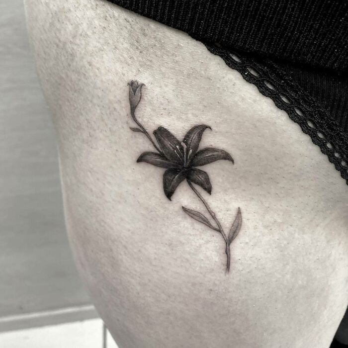 Black flower tattoo on skin, resembling a blossoming lily with a stem and bud.