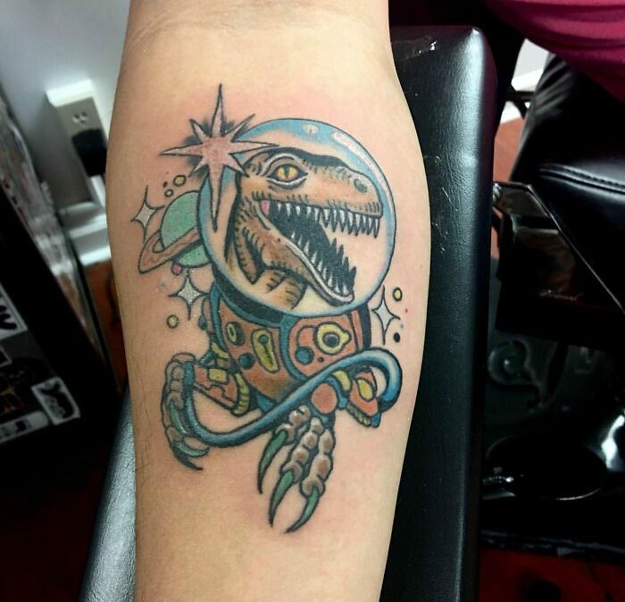 A colorful space tattoo featuring a dinosaur in an astronaut suit on a forearm.