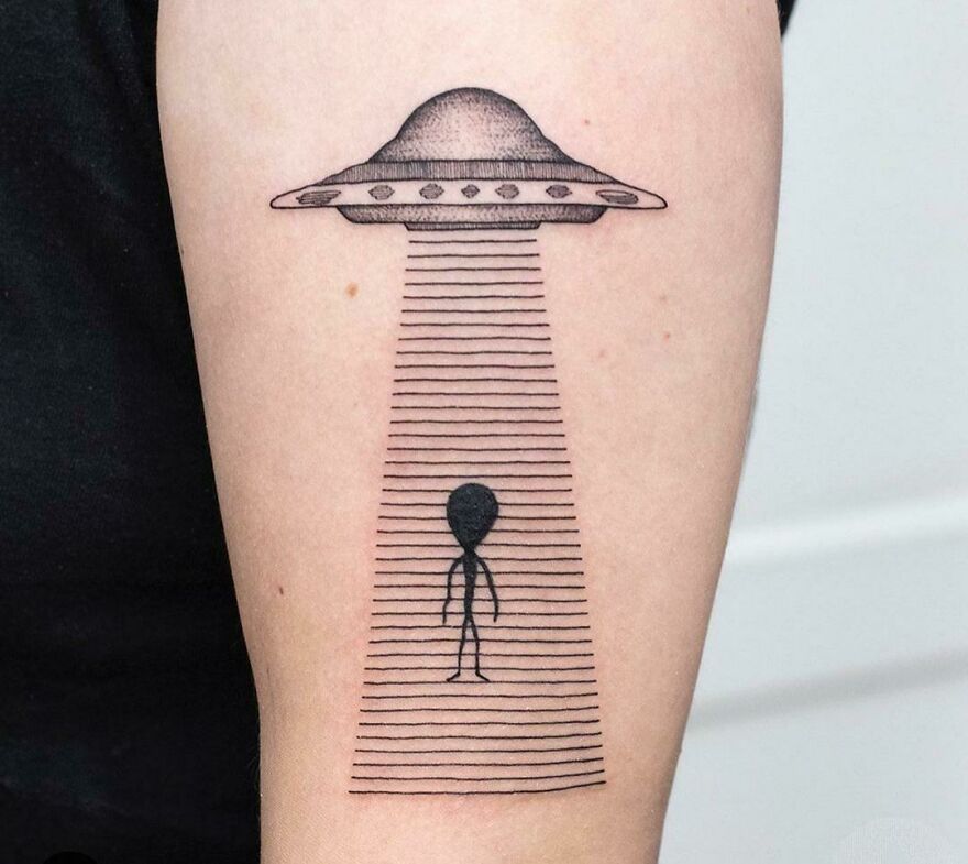 Alien and UFO space tattoo on forearm, featuring a spacecraft beaming up a stick-figure alien illustration.
