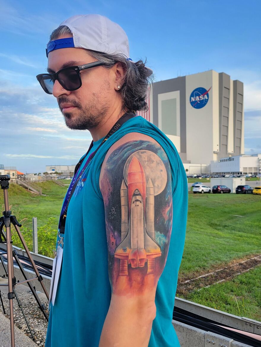 Man with space tattoo of a shuttle and moon on his arm, standing near NASA facility, showcasing outer space art.