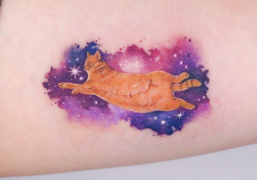 Tattoo of an orange cat floating in a galaxy-themed background, featuring cool space design elements.