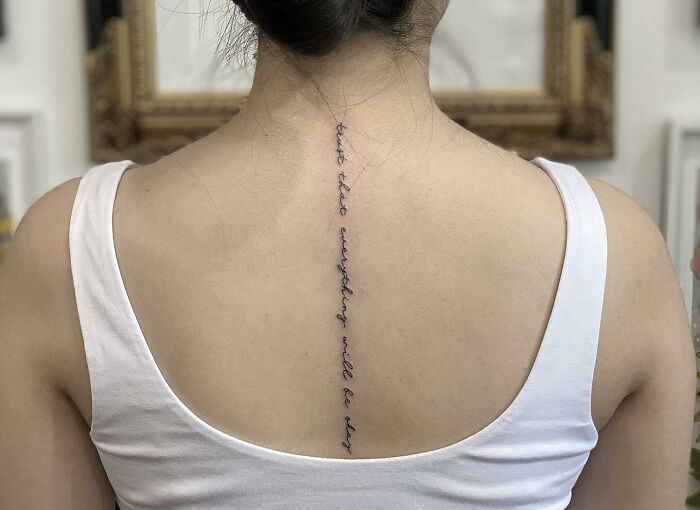 Back tattoo with script in a vertical line, embodying self-love and empowerment, on person wearing a white tank top.