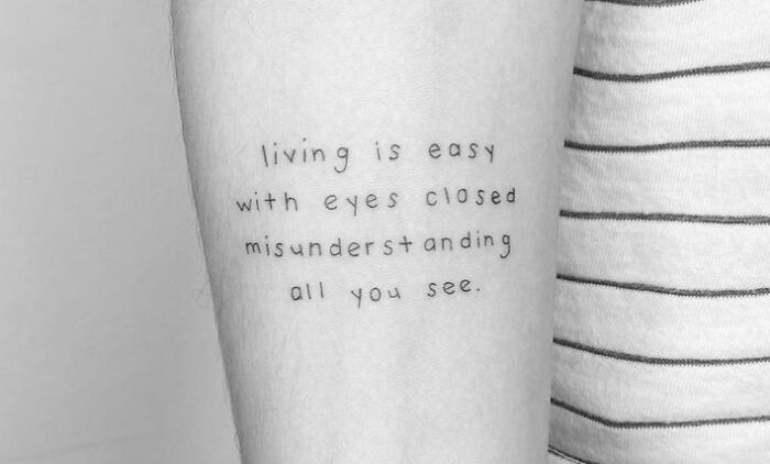 Minimalist self-love tattoo with the text "living is easy with eyes closed misunderstanding all you see" on an arm.