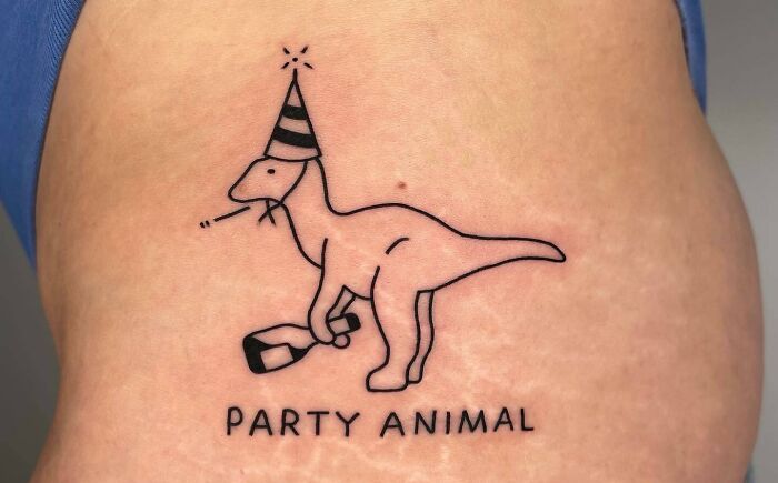Self-love tattoo featuring a grumpy dinosaur with a party hat, captioned "Party Animal."