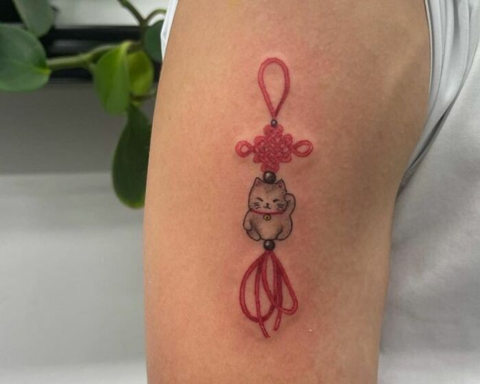 Red luck cat pendant tattoo on arm, symbolizing self-love and good fortune.