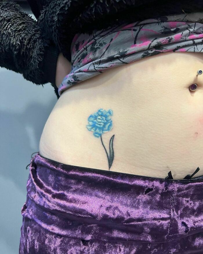 Blue flower tattoo on skin, resembling a blossom, with a purple velvet garment partially visible.