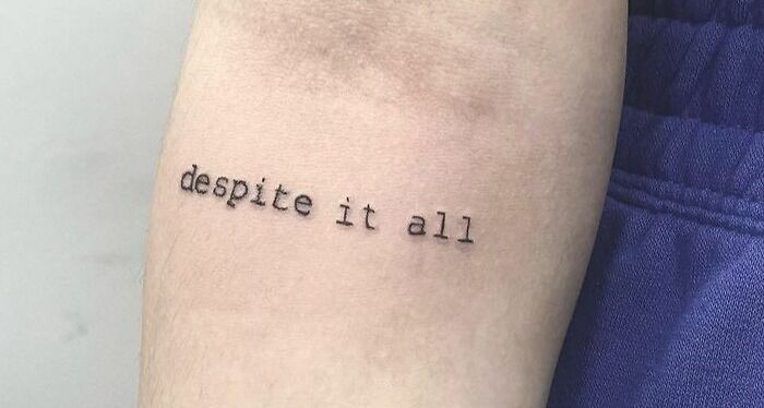 "Despite It All" self-love tattoo on forearm in simple black font.