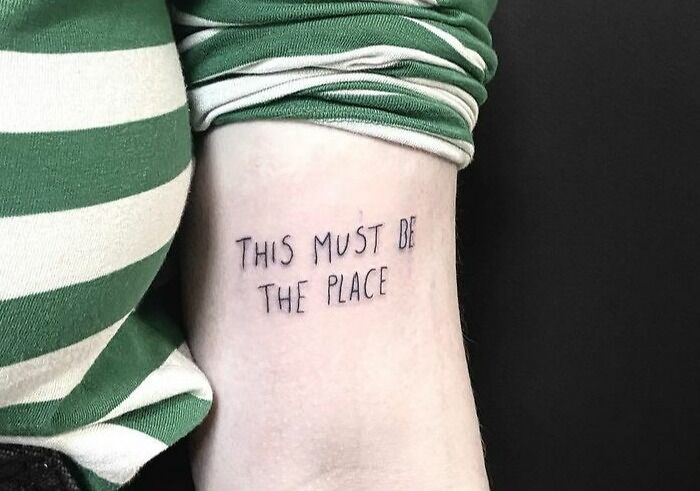 Close-up of a self-love tattoo reading "This Must Be the Place" on an arm wearing a striped green and white shirt.