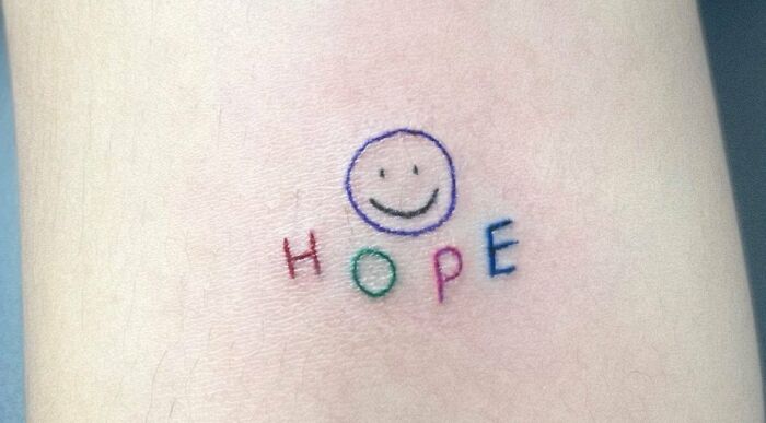 Minimalist self-love tattoo with a smiley face and the word "hope" in colorful letters.