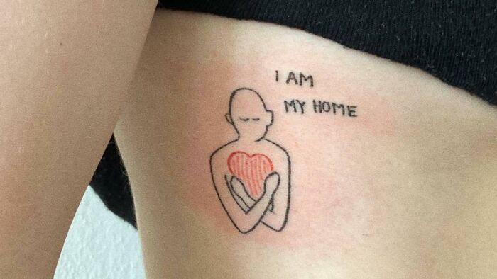 Self-love tattoo of person hugging a heart with text "I Am My Home" celebrating personal importance.