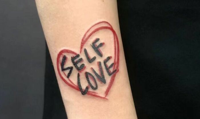 Heart-shaped self-love tattoo in red and black ink on a forearm.