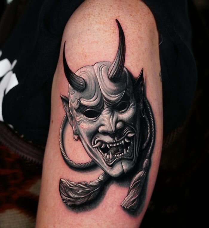 Realistic tattoo of a Japanese demon mask on an arm, showcasing detailed shadows and textures.
