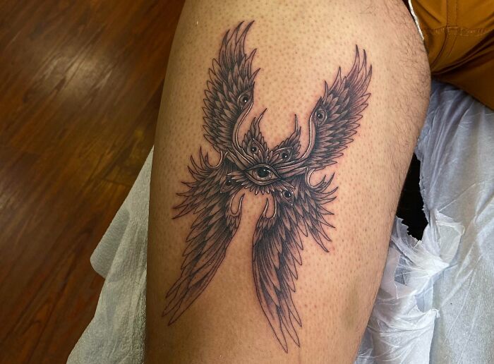 Realistic tattoo of a detailed angel design with a central eye, inked on a person’s thigh.