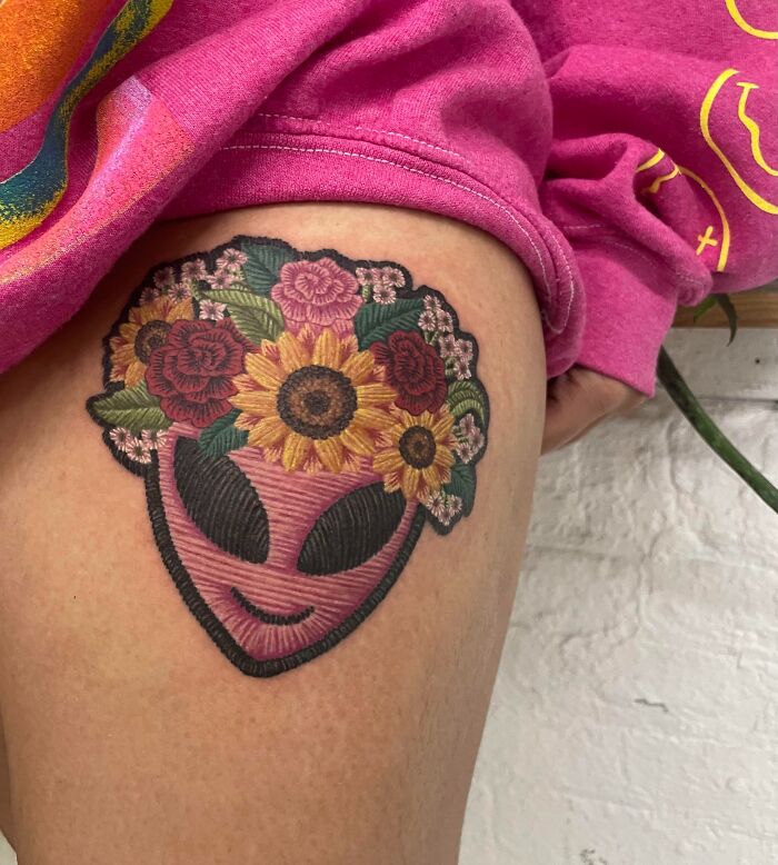 Realistic tattoo of an alien face with colorful flowers on a person's thigh, showcasing intricate design.