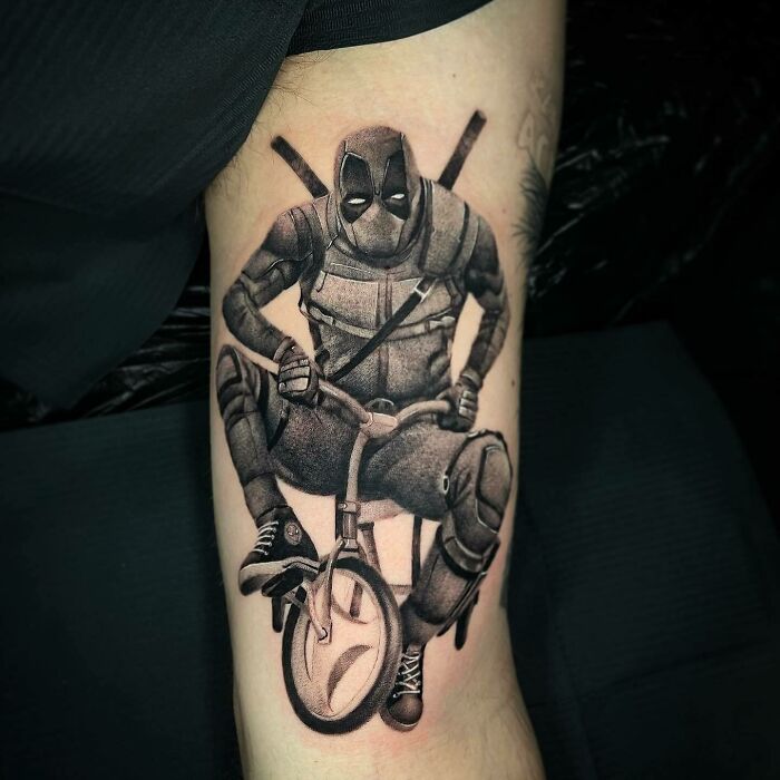 Marvel tattoo of Deadpool riding a small bicycle on a person's arm.