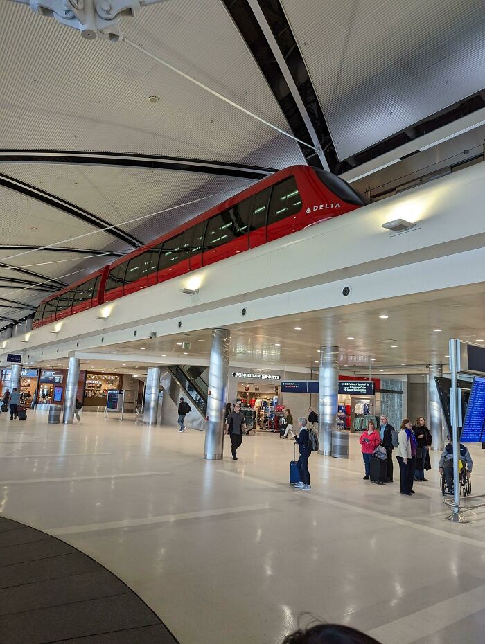 Red elevated train inside a spacious airport terminal with people walking and waiting, showcasing well-planned infrastructure.