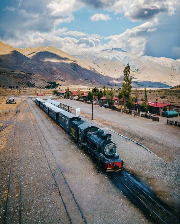 Vintage train running through a well-planned infrastructure station with mountains and cloudy sky in the background.