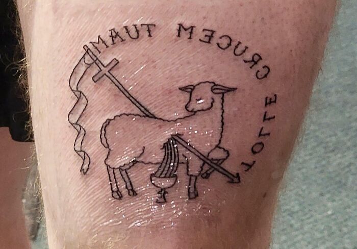 Word tattoo featuring a lamb, a cross, and the words "CRUCEM TUAM" and "TOLLE" on skin.