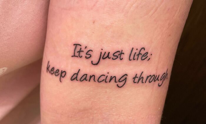Word tattoo on forearm reading "It's just life; keep dancing through" in black ink.