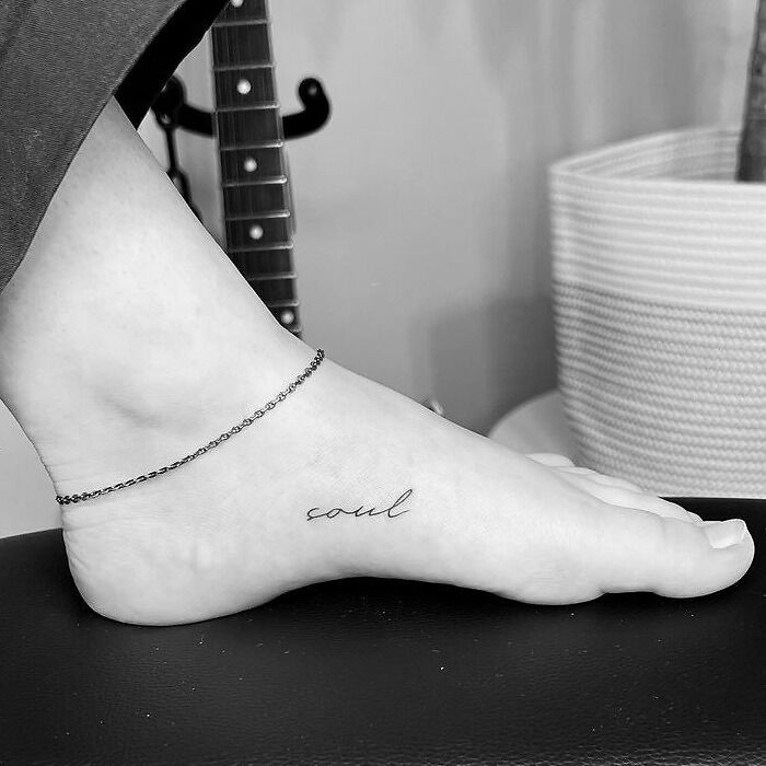 Word tattoo reading "soul" on a person's foot, minimalist design, next to a guitar and a plant.