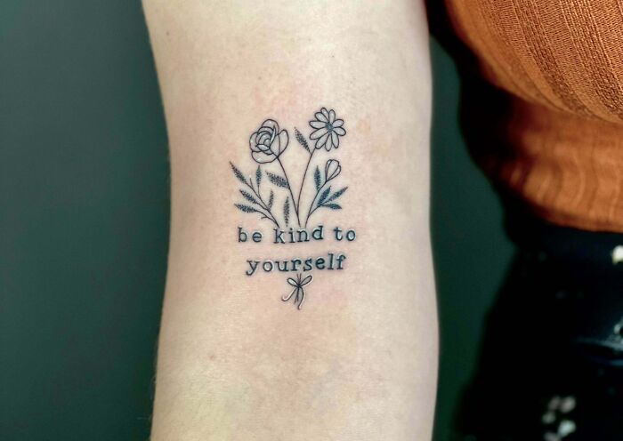 Word tattoo featuring flowers and the phrase "be kind to yourself" on an arm.