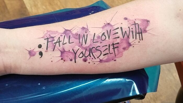 Tattoo with the phrase "Fall in love with yourself" on forearm, featuring purple watercolor background.