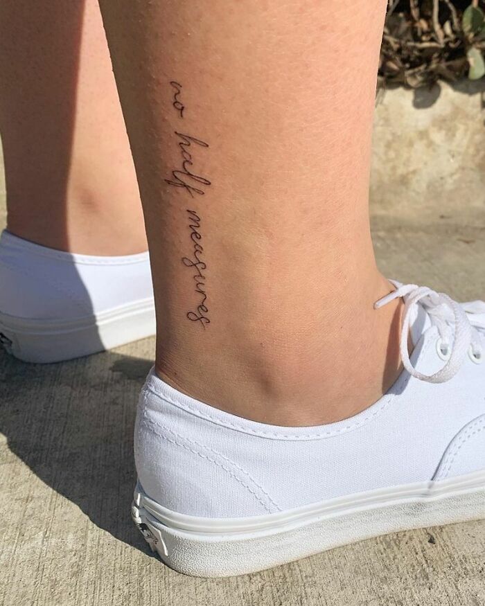 Minimalist word tattoo on leg, reading "no half measures," shown above white sneakers.