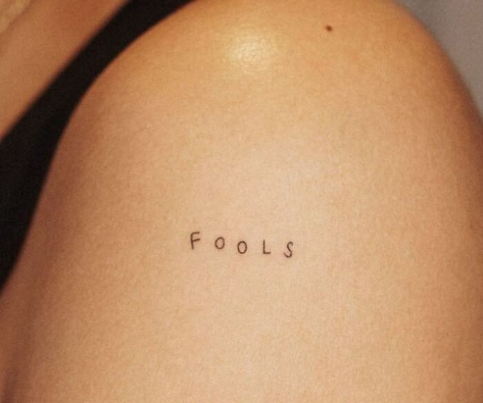 Small "FOOLS" word tattoo on upper arm.