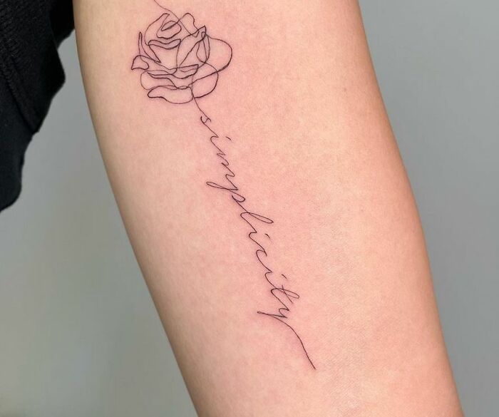 Minimalist word tattoo with "simplicity" forming the stem of a rose, inked on an arm.