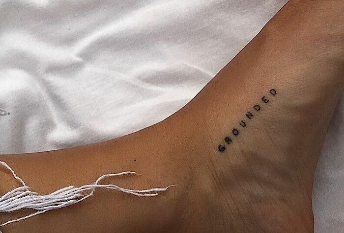 "Word tattoo on ankle, text reads 'grounded' in black ink. Simple design for new ink inspiration."