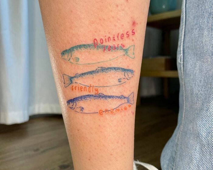 Tattoo of three fish with text: "pointless," "friendly," "enemy" on a person's leg. Word tattoo idea inspiration.