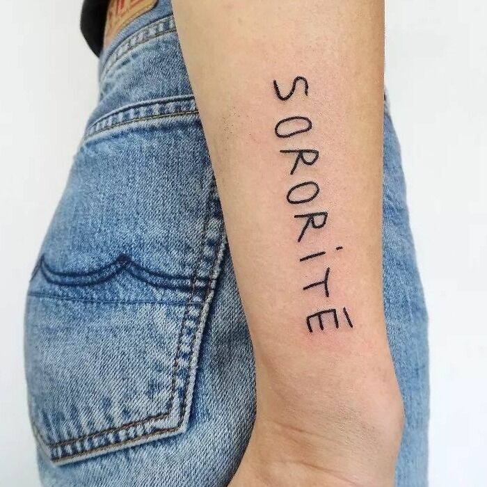 Close-up of an arm with "sororité" word tattoo in simple black font next to blue jeans.