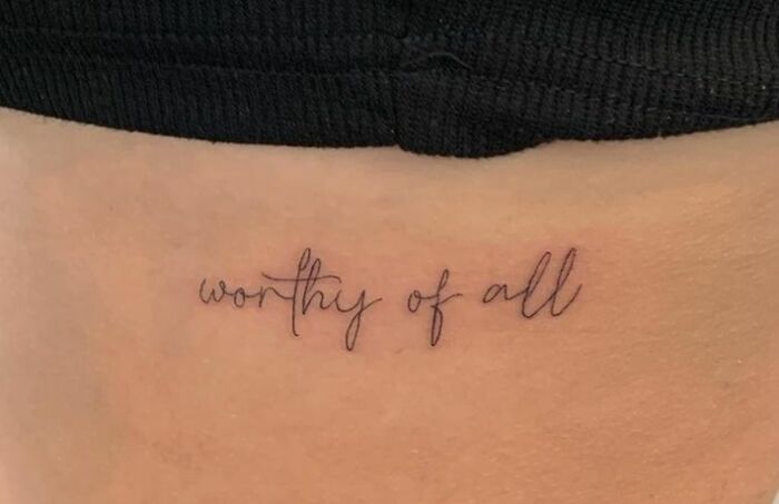 "Cursive word tattoo reading 'worthy of all' on skin."