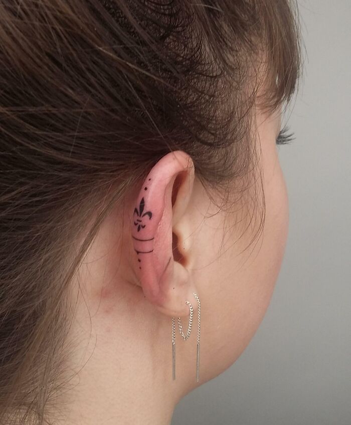 Ear tattoo with a fleur-de-lis design, featuring subtle black ink detail behind a woman's ear, paired with dangling earrings.